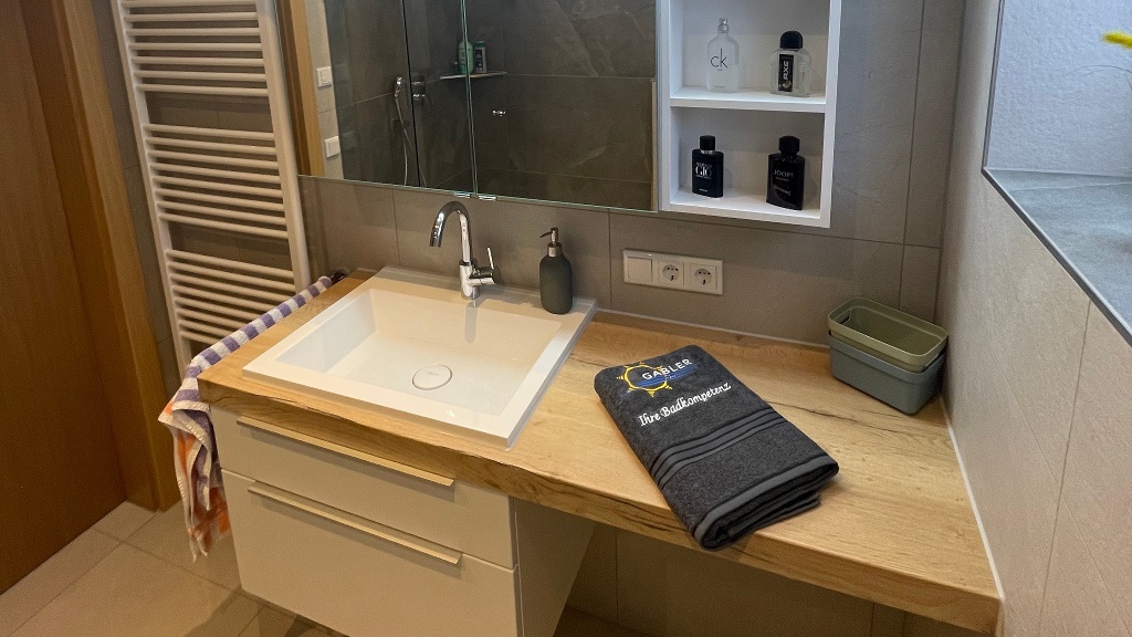 In einem Haus, das viele Generationen und Umbauten erlebt hat, entschied sich unsere Kundschaft, das Erdgeschoss neu zu gestalten. Die Herausforderung lag insbesondere im Badezimmer, da diese Räume früher eher klein gestaltet wurden. Zusätzlich gab es einen engen Raum nebenan für die Waschmaschine, der wenig Platz für Bewegung und Abstellfläche bot. 

Nach eingehender Prüfung entschieden wir uns schnell für eine Raumerweiterung. Aufgrund der veralteten Rohrleitungen führten wir eine Komplettbadsanierung durch, inklusive neuer Trink- und Abwasserleitungen. Dadurch konnten wir nicht nur eine bodengleiche Dusche und eine großzügige Waschtischanlage von Laguna Badwelten planen, sondern auch das Bad mit einem WC ausstatten. 

Die Waschtischanlage wurde nach den Wünschen der Kundschaft geplant und installiert, wodurch auch die Waschmaschine problemlos im Badezimmer Platz fand. In Zusammenarbeit mit einem Glaser konnten wir die Glaswandlösung – eine Glaswand mit Pendeltür – optimal nutzen, wenn nicht geduscht wird.