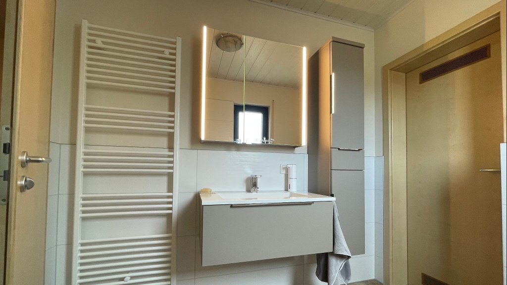 Trotz der beschränkten Fläche von nur 5 m² konnten wir im Badezimmer die Wünsche unserer Kundschaft erfüllen. Unsere Kunden freuten sich über eine geräumige, bodengleiche Dusche, die mit einer Duscharmatur inklusive Regenkopfbrause ausgestattet wurde. Die Duschabtrennung – eine Glaswand mit Pendeltür – wurde ebenfalls installiert. 

Die Möglichkeit, die Dusche einzuklappen und somit mehr Raum zu schaffen, erweist sich als großer Vorteil. Die Waschtischanlage von Laguna Badwelten fand ebenfalls ihren Platz im Bad. Statt einer herkömmlichen Armatur wurde eine elektronische Mischerarmatur von Hansa montiert. Dank Infrarotsensoren erkennt die Armatur, wenn jemand seine Hände waschen möchte, wodurch das lästige Spritzen beim Schließen des Wasserhahns vermieden wird. 

Eine weitere Besonderheit in diesem Bad ist das WC, ein Dusch-WC von Vigour, das sich auf die wichtigste Eigenschaft konzentriert: die gründliche Reinigung mit Wasser. Perfekt für diejenigen, die ein Dusch-WC ohne überflüssigen Schnickschnack bevorzugen.