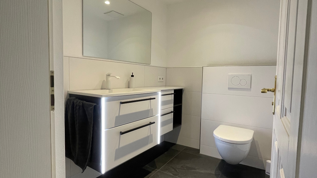 Neben dem Hauptbad wurde auch das Gäste-WC der Kundschaft in Greding modernisiert. Aufgrund der Möglichkeit, das WC zu versetzen, kam uns die Idee, eine größere Waschtischanlage von Laguna Badwelten in den Raum zu integrieren. Eine kleine Besonderheit für die Waschtischanlage haben wir ebenfalls umgesetzt. 

Die Beleuchtung direkt an der Waschtischanlage ist mit einem Bewegungsmelder ausgestattet. Somit erlebt die Kundschaft ein angenehmes Nachtlicht, wenn man am Abend noch die Toilette benutzt. Auch im Gäste-WC sind die Farben Weiß & Anthrazit präsent. 

Die Serie Vivenis von Hansgrohe in Weiß matt fügt sich nahtlos in das Konzept ein. Die großflächigen Fliesen, die in beiden Räumen verwendet wurden, verleihen dem Raum ein größeres Erscheinungsbild.